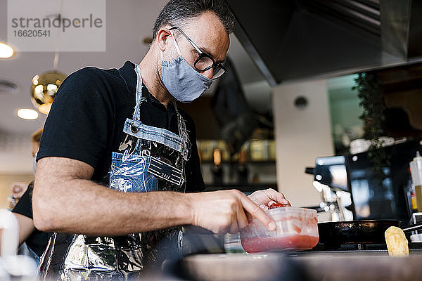 Männlicher Barista mit Gesichtsmaske bei der Zubereitung von Speisen in einem Café während des COVID-19-Ausbruchs