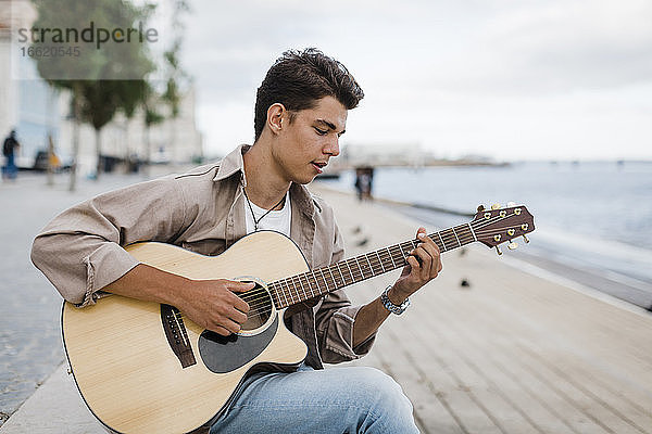 Junger Mann spielt Gitarre beim Üben an der Promenade gegen den Himmel