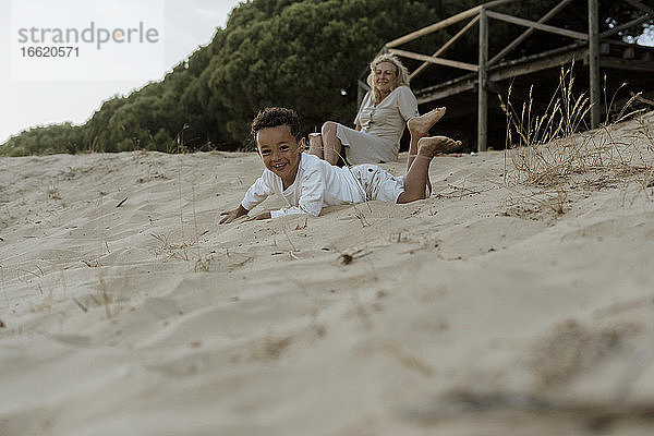 Großmutter sitzt  während der Enkel am Strand im Sand liegt