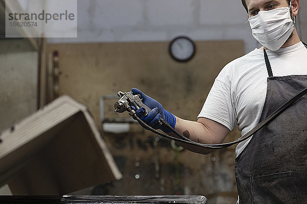 Mann mit Gesichtsmaske  der mit einer Spritzpistole auf Holz spritzt  während er in einer Fabrik steht