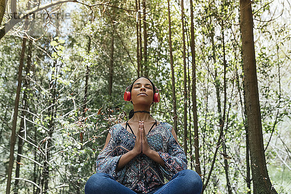 Junge Frau mit geschlossenen Augen  die über Kopfhörer Musik hört  während sie im Park gegen Bäume meditiert
