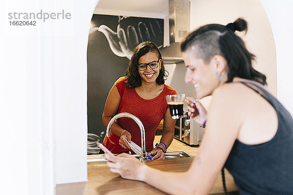 Lächelnde junge Frau beim Geschirrspülen  während ihr Freund Kaffee trinkt und eine SMS auf seinem Smartphone schreibt