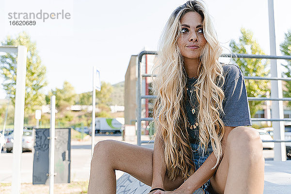 Nachdenkliche Frau mit langen blonden Haaren  die wegschaut  während sie im Skateboardpark sitzt