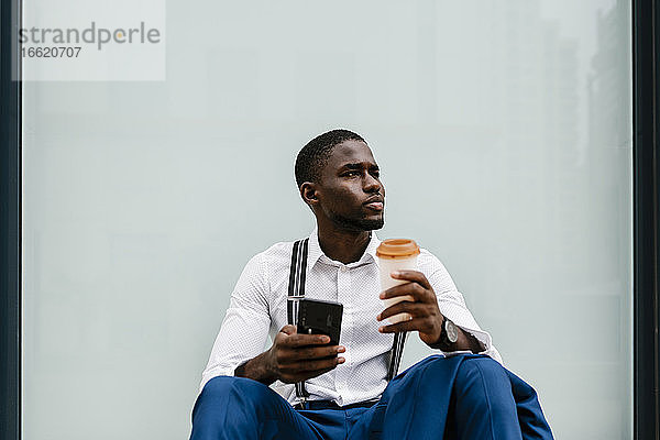 Nachdenklicher männlicher Unternehmer  der ein Smartphone und einen wiederverwendbaren Becher in der Hand hält  während er in der Stadt am Fenster sitzt