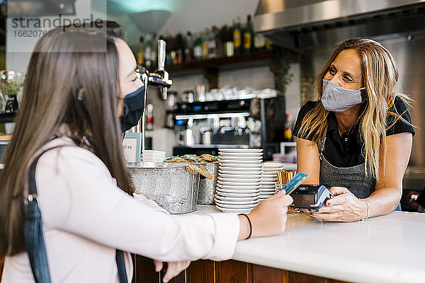 Geschäftsfrau mit Gesichtsmaske  die während der COVID-19-Krise am Schalter eines Cafés mit Kreditkarte bezahlt