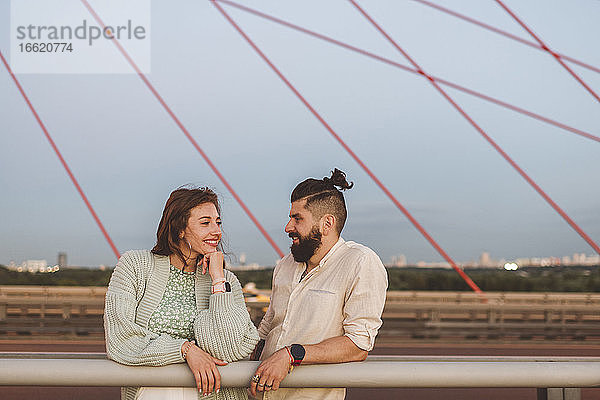 Lächelnder Freund und Freundin stehen auf einer Brücke gegen den Himmel in der Stadt bei Sonnenuntergang
