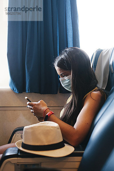 Mittlere erwachsene Frau mit Maske  die ein Mobiltelefon benutzt  während sie auf einem Sitz in einem Kreuzfahrtschiff sitzt