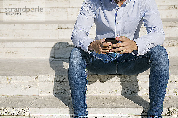 Mann surft im Internet über sein Smartphone  während er an einem sonnigen Tag auf einer Treppe sitzt