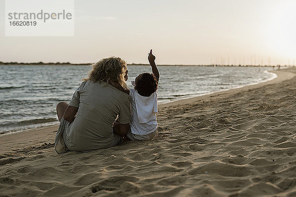 Enkel  der in den Himmel zeigt  während er mit seiner Großmutter am Strand sitzt  während die Sonne untergeht