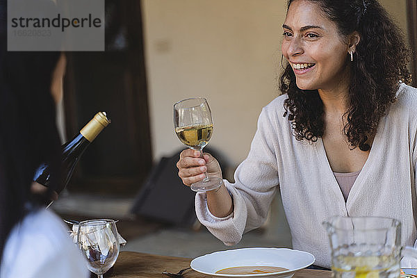 Lächelnde Frau hält ein Weinglas in der Hand und sieht einen Freund an  der am Esstisch im Hinterhof sitzt