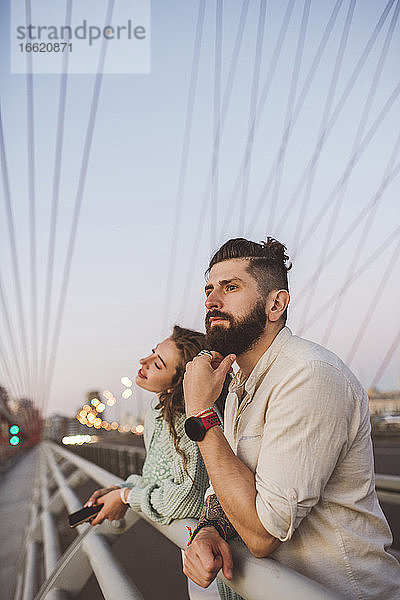 Nachdenkliches Paar  das sich bei Sonnenuntergang auf das Geländer einer Brücke in der Stadt stützt