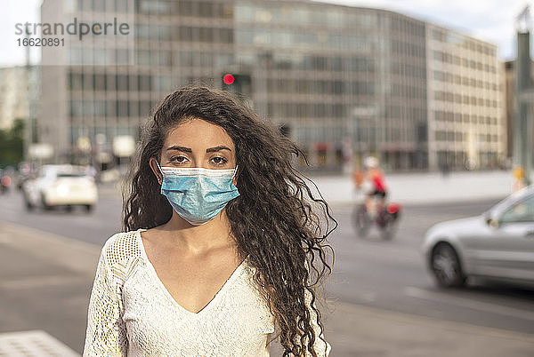 Junge Frau mit gewelltem Haar trägt eine Maske  während sie auf der Straße in der Stadt steht