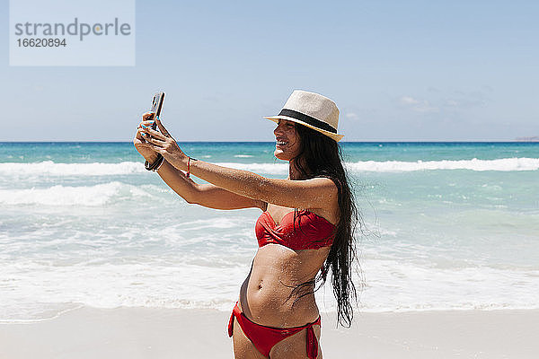 Frau im Bikini macht ein Selfie mit ihrem Smartphone  während sie am Meer steht