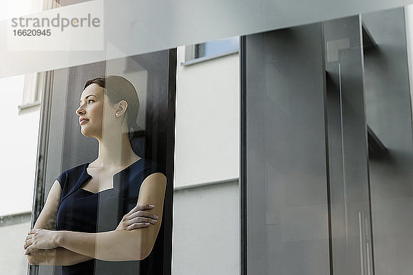 Nachdenkliche Unternehmerin mit verschränkten Armen im Büro stehend  gesehen durch eine Glastür