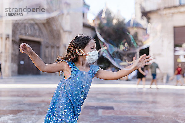 Unbekümmertes Mädchen mit Maske läuft auf der Straße in der Stadt und spielt mit einer Seifenblase