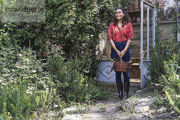 Lächelnde reife Frau schaut weg und hält einen Korb  während sie im Hinterhof steht