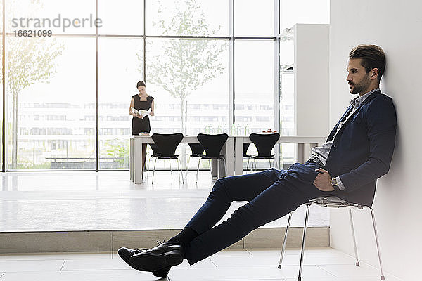 Nachdenklicher Geschäftsmann auf einem Stuhl sitzend  während eine Kollegin im Hintergrund im Büro arbeitet