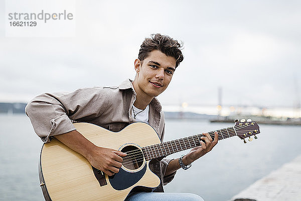 Hübscher junger Mann spielt Gitarre beim Üben an der Promenade gegen den Himmel