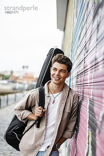 Lächelnder junger Mann  der wegschaut  während er eine Gitarre hält und sich an einen Fensterladen mit Graffiti lehnt
