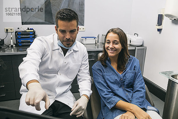 Ein männlicher Zahnarzt erklärt einer lächelnden jungen Patientin über einem Laptop in einer Klinik ein medizinisches Verfahren