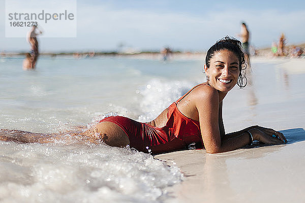 Lächelnde erwachsene Frau in Dessous am Strand liegend gegen den Himmel