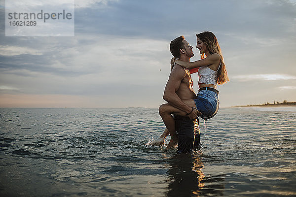 Mann trägt Frau während des Sonnenuntergangs am Strand im Wasser stehend