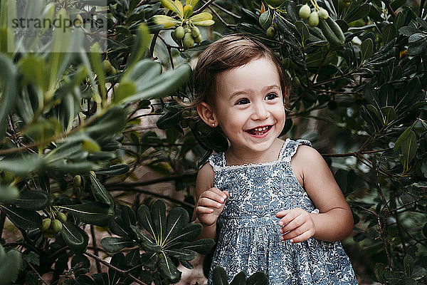 Glückliches kleines Mädchen  das in einem öffentlichen Park vor grünen Blättern steht