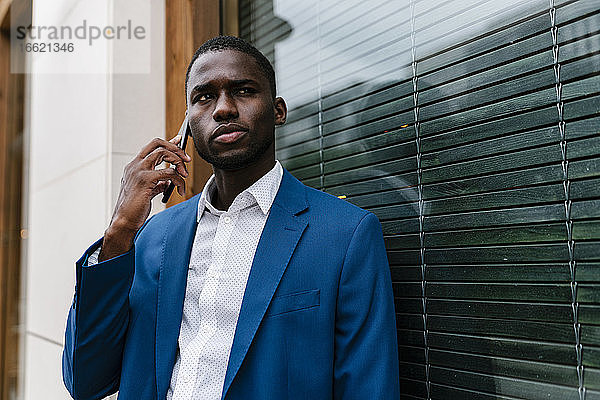 Geschäftsmann  der wegschaut  während er während eines Anrufs durch sein Mobiltelefon lauscht und sich an ein Fenster lehnt