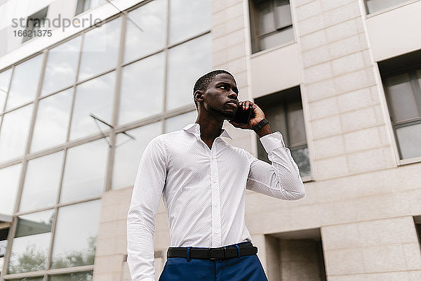 Männlicher Berufstätiger am Telefon  der wegschaut  während er vor einem Bürogebäude in der Stadt steht