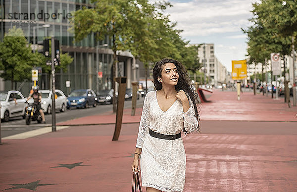 Junge Frau in weißem Kleid schaut weg  während sie auf einem Fußweg in der Stadt geht