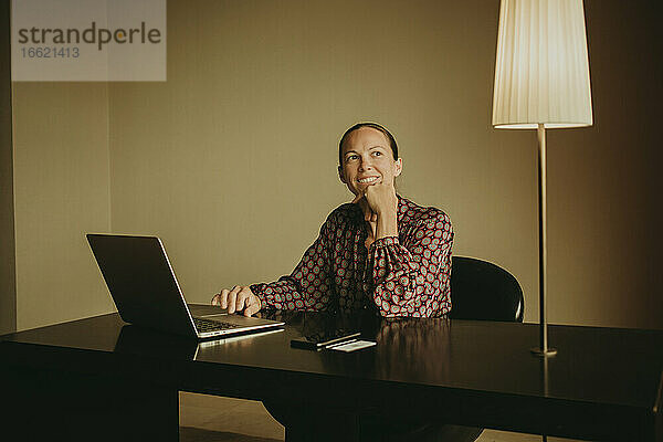 Lächelnde Unternehmerin  die bei der Arbeit am Laptop im Büro wegschaut