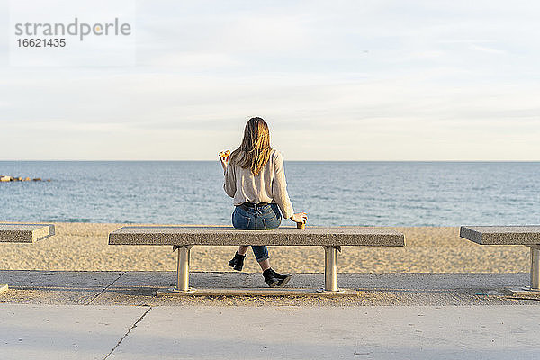 Junge Frau schaut auf das Meer  während sie auf einer Bank sitzt und den Sonnenuntergang genießt