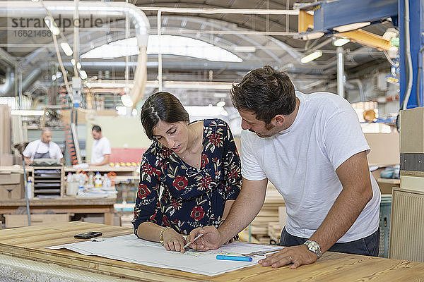 Projektleiter und Architekt besprechen den Plan mit einem Mitarbeiter  der im Hintergrund in einer Fabrik arbeitet