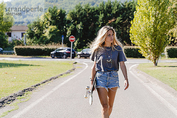 Schöne junge blonde Frau schaut weg  während sie ein Skateboard hält und auf der Straße in der Stadt an einem sonnigen Tag läuft