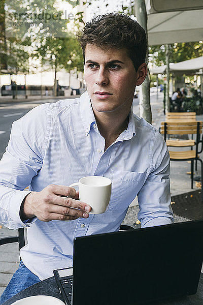 Nachdenklicher junger Mann hält eine Kaffeetasse  während er mit seinem Laptop in einem Straßencafé sitzt