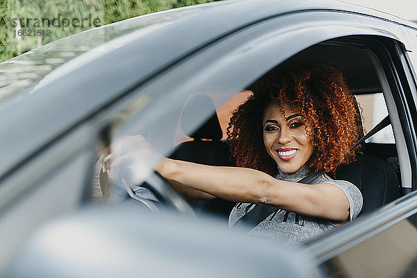 Lächelnde junge Frau mit Afrohaar am Steuer eines Autos  gesehen durch ein Fenster