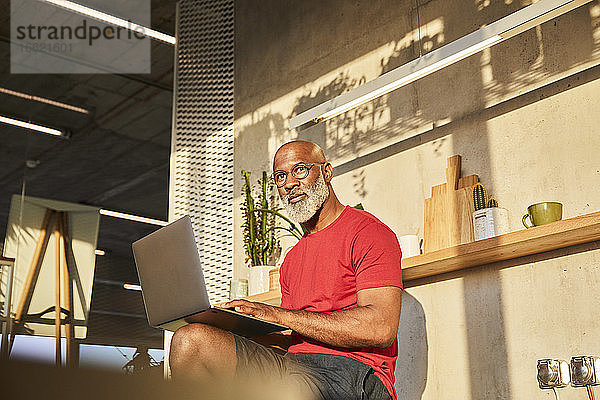 Reifer Mann schaut weg  während er zu Hause an einem sonnigen Tag einen Laptop benutzt