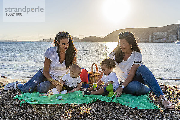 Schwestern mit Baby Söhne sitzen am Strand gegen den Himmel bei Sonnenuntergang