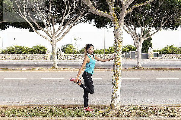 Junge lateinamerikanische Frau streckt ihr Bein aus und lehnt sich an einen Baumstamm an der Straße
