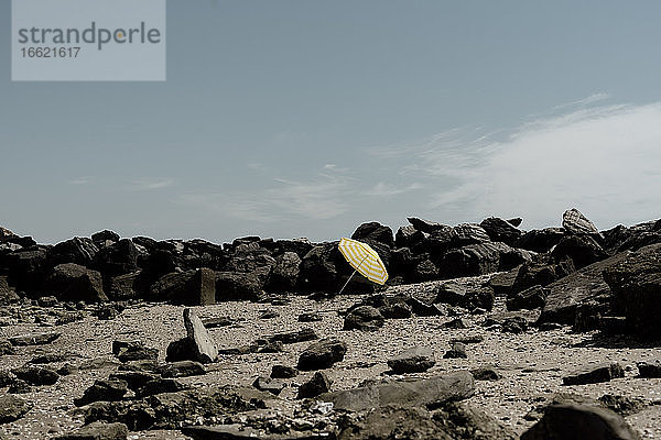 Gelber Regenschirm auf Sand inmitten von Felsen am Strand gegen den Himmel an einem sonnigen Tag