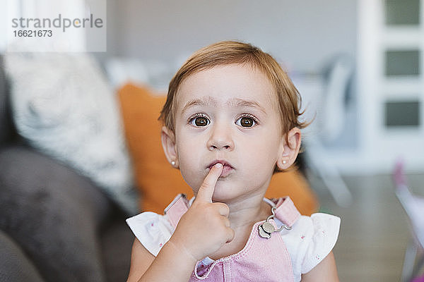 Nachdenkliches kleines Mädchen mit Finger auf den Lippen  das zu Hause auf dem Sofa sitzt