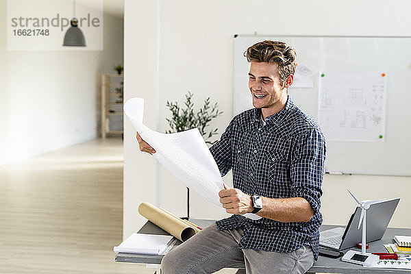 Lächelnder junger Mann  der eine Blaupause hält und auf einem Schreibtisch im Büro sitzt