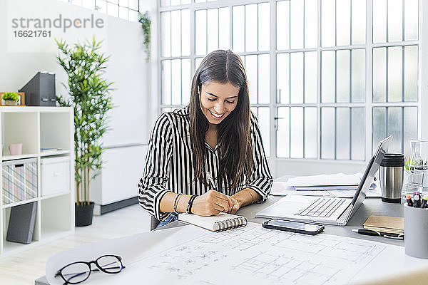 Lächelnde junge Architektin  die am Schreibtisch im Büro sitzt und in ihr Tagebuch schreibt