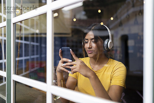 Schöne junge Frau hört Musik und benutzt ihr Smartphone in einer Bar  gesehen durch ein Fenster