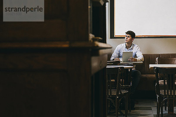Nachdenklicher Mann  der eine Kaffeetasse und ein digitales Tablet hält  während er in einem Café sitzt