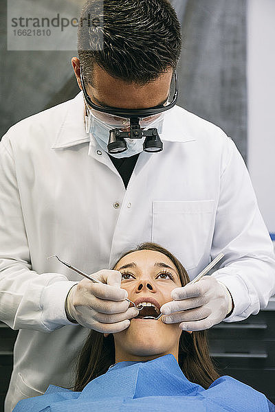 Männlicher Zahnarzt mit chirurgischer Lupenbrille bei der Untersuchung der Zähne einer jungen Patientin in der Klinik