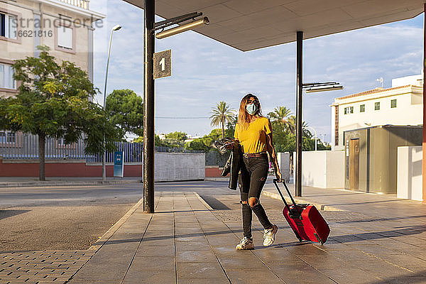 Junge Frau mit Schutzmaske beim Gehen mit Gepäck an der Bushaltestelle