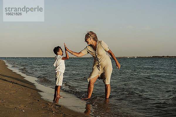 Glückliche Großmutter und Enkel machen High Five zueinander am Strand während des Sonnenuntergangs