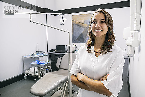 Lächelnde junge Zahnarzthelferin steht mit verschränkten Armen in einer Klinik