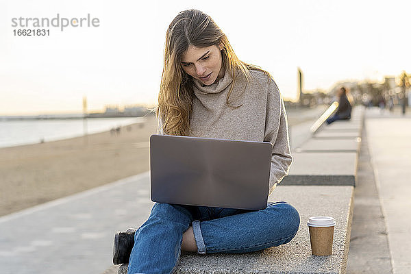 Schöne junge Frau  die einen Laptop benutzt und mit einem Einwegbecher auf einer Bank an der Strandpromenade gegen den Himmel sitzt